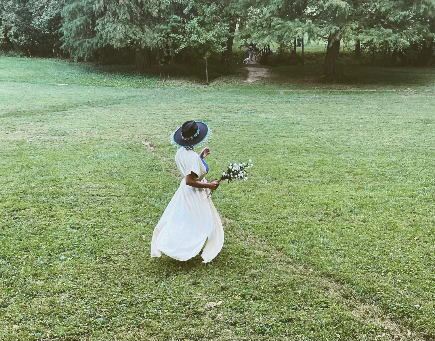 A figure in a flowing white dress and black brimmed hat running through a field of grass.