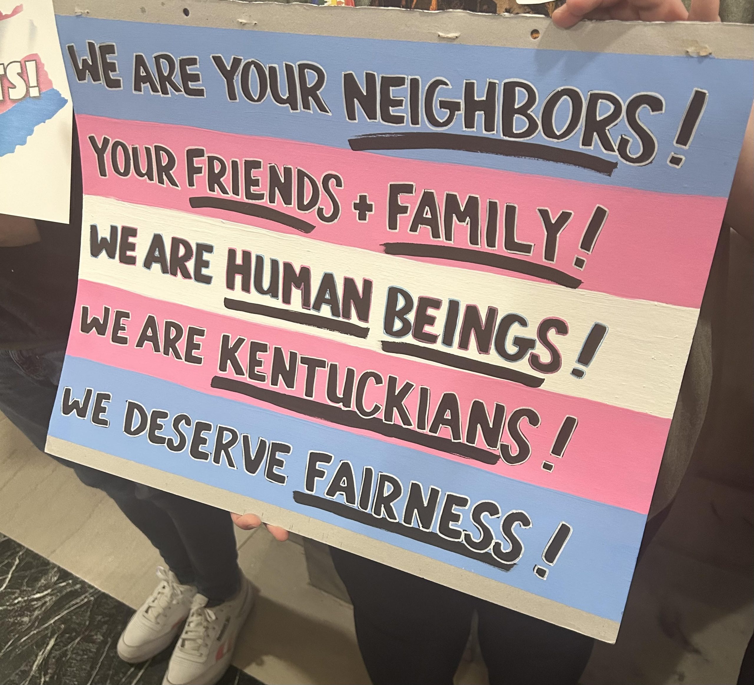 The image shoes a poster made of the colors of the trans pride flag and it reads "We are your neighbors, your friends and family, we are human beings, we are Kentucky, we deserve fairness"
