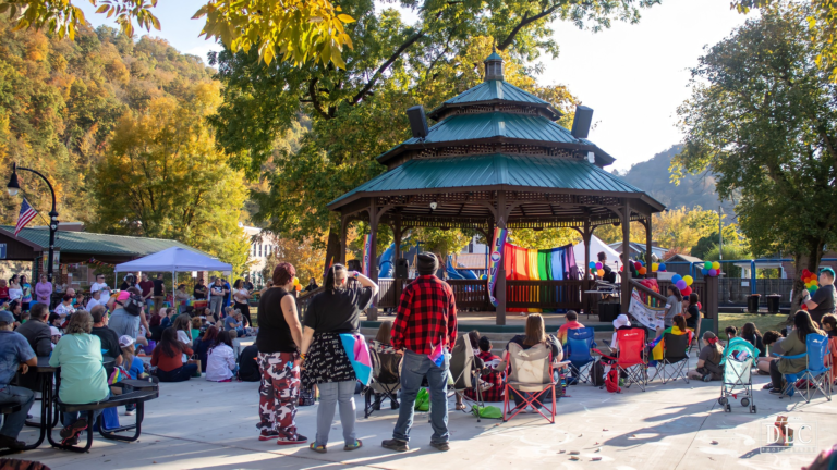 A crowd gathers around a gazebo with rainbow flags at Pikeville Pride, with mountains behind