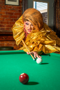 Drag performer Molly Mormen lines up a shot on a pool table at Rosie’s Tavern in Covington, Kentucky, (Northern Kentucky) wearing a gold ruffled gown and red jewelry.