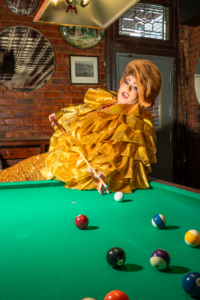 Drag performer Molly Mormen poses dramatically on a pool table in a shimmering gold gown at Rosie’s Tavern in Covington, Kentucky.