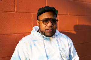 Xian Brooks, a Black transgender man from Louisville, Kentucky, wearing sunglasses, a black cap, and a tie-dye hoodie, stands against an orange brick wall in the afternoon sunlight.