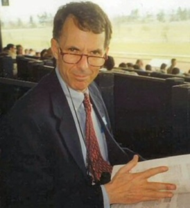 Judge Lewis Grant Paisley reads a document while seated in a courtroom gallery, wearing glasses, a suit, and a patterned tie.