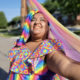 Sophia Harris smiles brightly outdoors while holding a flowing rainbow-colored fabric above her head. She wears a vibrant multicolored outfit and stands in sunlight on a neighborhood street.