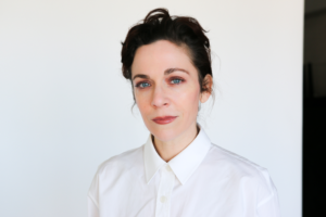 Actor and writer Jen Tullock poses against a plain white background wearing a crisp white button-down shirt. The Kentucky-born creator of Nothing Can Take You From the Hand of God and star of Apple TV’s Severance looks directly at the camera with a calm, confident expression.
