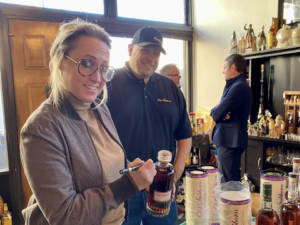 A woman signs a bourbon bottle at Revival Vintage Bottle Shop and Bar in Covington, Kentucky, while a staff member smiles beside her. Shelves of vintage spirits are visible in the background.