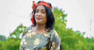 Zachee Imanitwitaho, a transgender woman from Louisville, Kentucky, wearing a gold floral dress and an orange headscarf, stands outdoors with trees in the background.