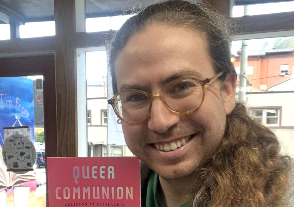 Davis Shoulders with long hair and glasses smiling indoors while holding up a pink book titled “Queer Communion: Religion in Appalachia.” Sunlight filters through the windows behind them, revealing part of a small-town street outside.