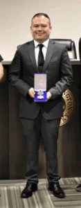 Covington police officer, Zachary Stayton, in a dark suit and tie stands indoors holding an award medal in a blue case, posing for a formal photo in front of a wooden wall with the Covington city seal partially visible behind him.