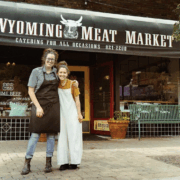 Reka’s Wives Shelbi and Ashley Nation, known as the Nation wives, stand arm in arm in front of Wyoming Meat Market. They are smiling, wearing work aprons, and standing on the brick sidewalk beneath the shop’s black awning that reads “Wyoming Meat Market.” A bench and storefront windows are visible behind them.