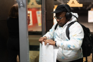 Xian Brooks, a Black transgender man from Louisville, Kentucky, wearing headphones and a tie-dye hoodie, examines a paper target at an indoor gun range.