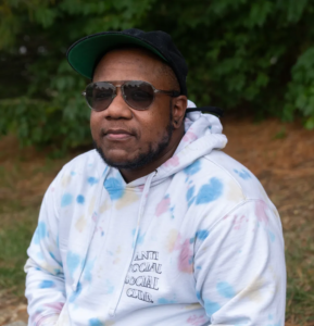 Xian Brooks, a Black transgender man from Louisville, Kentucky, wearing sunglasses, a black cap, and a tie-dye hoodie that reads “Anti Social Social Club,” sits outdoors with greenery in the background.