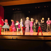 Slay Bells A lineup of drag performers stands across a brightly lit stage at the Slay Bells holiday show, wearing festive gowns, wigs, and glamorous costumes. Large red letters spell out “IVA” in front of them, with oversized Christmas ornaments decorating the background. Red curtains frame the stage as the performers pose and interact during the fundraiser for Moveable Feast Lexington.