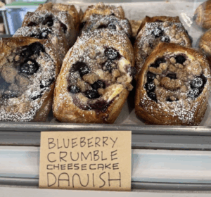 Tray of blueberry crumble cheesecake danishes topped with powdered sugar at a bakery.