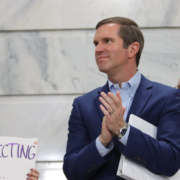 485652387_1053658180125129_91627383097881937_n Gov. Andy Beshear, in a blue suit claps while standing inside a marble government building during a Fairness Rally, as people around him hold protest signs advocating for LGBTQ+ protections.