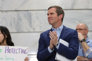 Gov. Andy Beshear, in a blue suit claps while standing inside a marble government building during a Fairness Rally, as people around him hold protest signs advocating for LGBTQ+ protections.