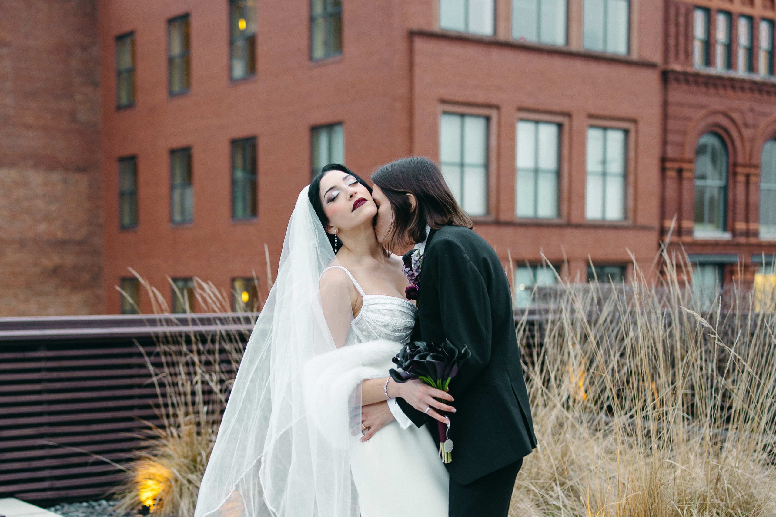 Heather Shaw and Lilly Jean Coiner embrace during their wedding portraits outdoors, with Coiner wearing a white wedding gown and veil and Shaw wearing a black suit while holding a bouquet of dark flowers. Brick buildings and tall grasses are visible in the background.