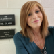 Dr. Bobbie Glass with shoulder-length light brown hair takes a selfie in a school hallway. She is wearing a teal top and layered necklaces. Behind her on a cinderblock wall is a black sign reading “106 Classroom Dr. Glass” with Braille beneath it, and a plaque that says “A Gift by WHAS Crusade for Children.”
