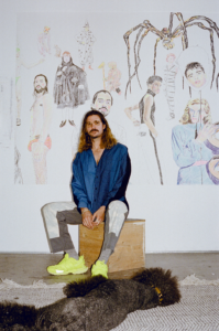 Artist John Brooks seated in front of large figurative drawings, surrounded by mixed-media artwork exploring memory, identity, and queer themes.