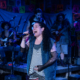 A performer sings passionately into a handheld microphone on a small stage under blue lighting, backed by a live band with guitars and drums. Colorful posters hang behind the musicians, creating a vibrant, community theater atmosphere.