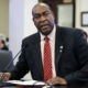 Sen. Donald Douglas, R-Nicholasville, speaks at a microphone during a Kentucky Senate Committee on Health Services meeting in Frankfort, presenting Senate Bill 72 (SB 72). He sits at a desk with papers in front of him, wearing a dark suit and red patterned tie, as attendees listen in the background.