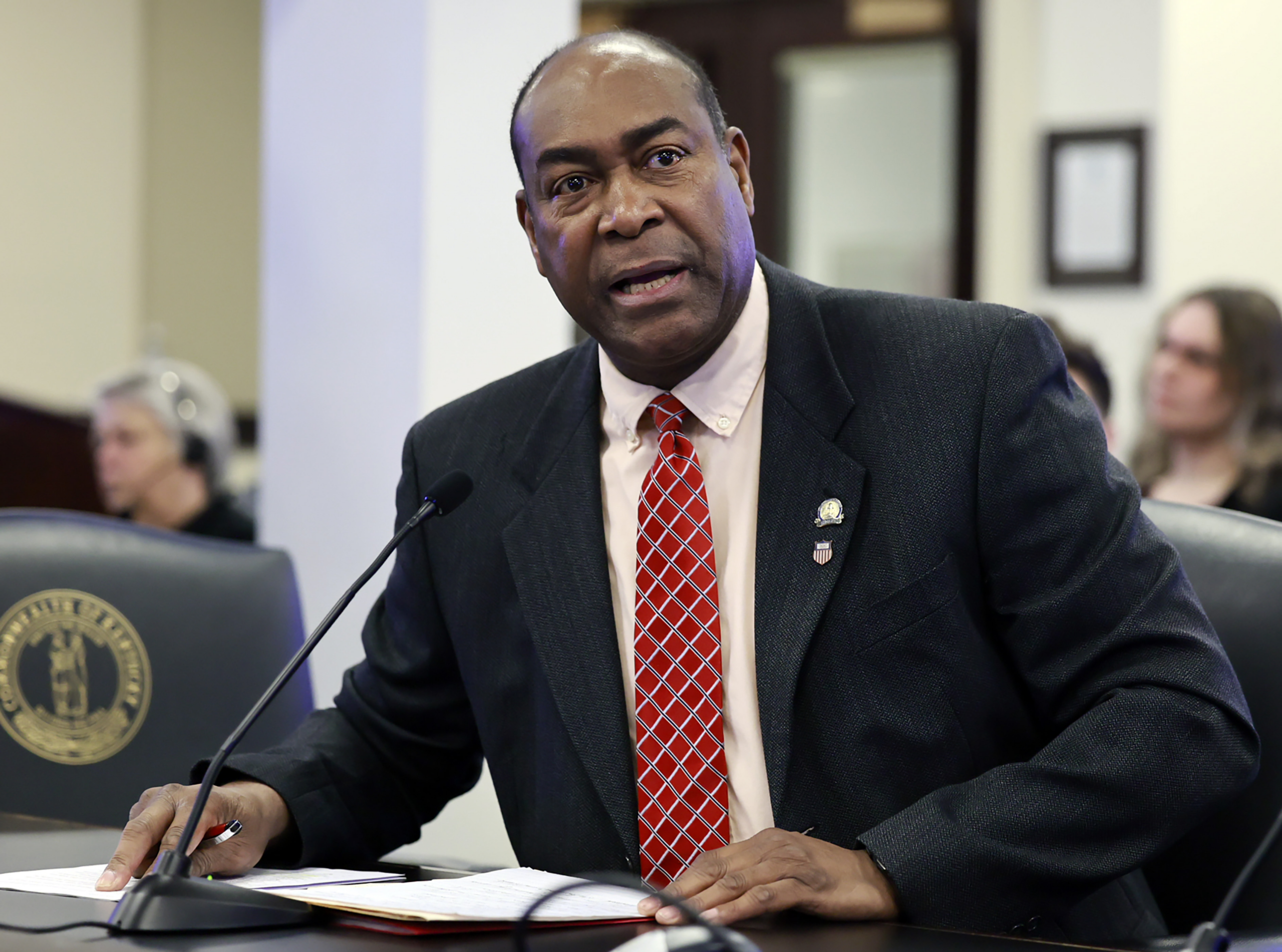 Sen. Donald Douglas, R-Nicholasville, speaks at a microphone during a Kentucky Senate Committee on Health Services meeting in Frankfort, presenting Senate Bill 72 (SB 72). He sits at a desk with papers in front of him, wearing a dark suit and red patterned tie, as attendees listen in the background.