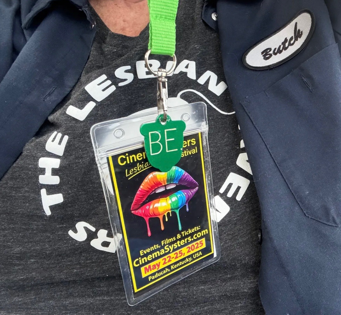 A close-up of a Cinema Systers Film Festival badge on a green lanyard featuring the rainbow lips logo and dates May 22–25, 2025, worn over a "Save the Lesbians" t-shirt with a "Butch" name patch. Description: A Cinema Systers 2025 festival badge hangs from the signature green lanyard, displaying the rainbow lips logo and website CinemaSysters.com. The attendee's "Butch" name patch and "Save the Lesbians" shirt perfectly capture the joyful, unapologetic spirit of the Cinema Systers community in Paducah, Kentucky.