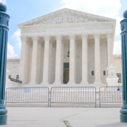 The U.S. Supreme Court building in Washington, D.C., framed between two dark green bollards, with metal barricades and an 'Area Closed' sign across the front steps