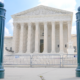 The U.S. Supreme Court building in Washington, D.C., framed between two dark green bollards, with metal barricades and an 'Area Closed' sign across the front steps