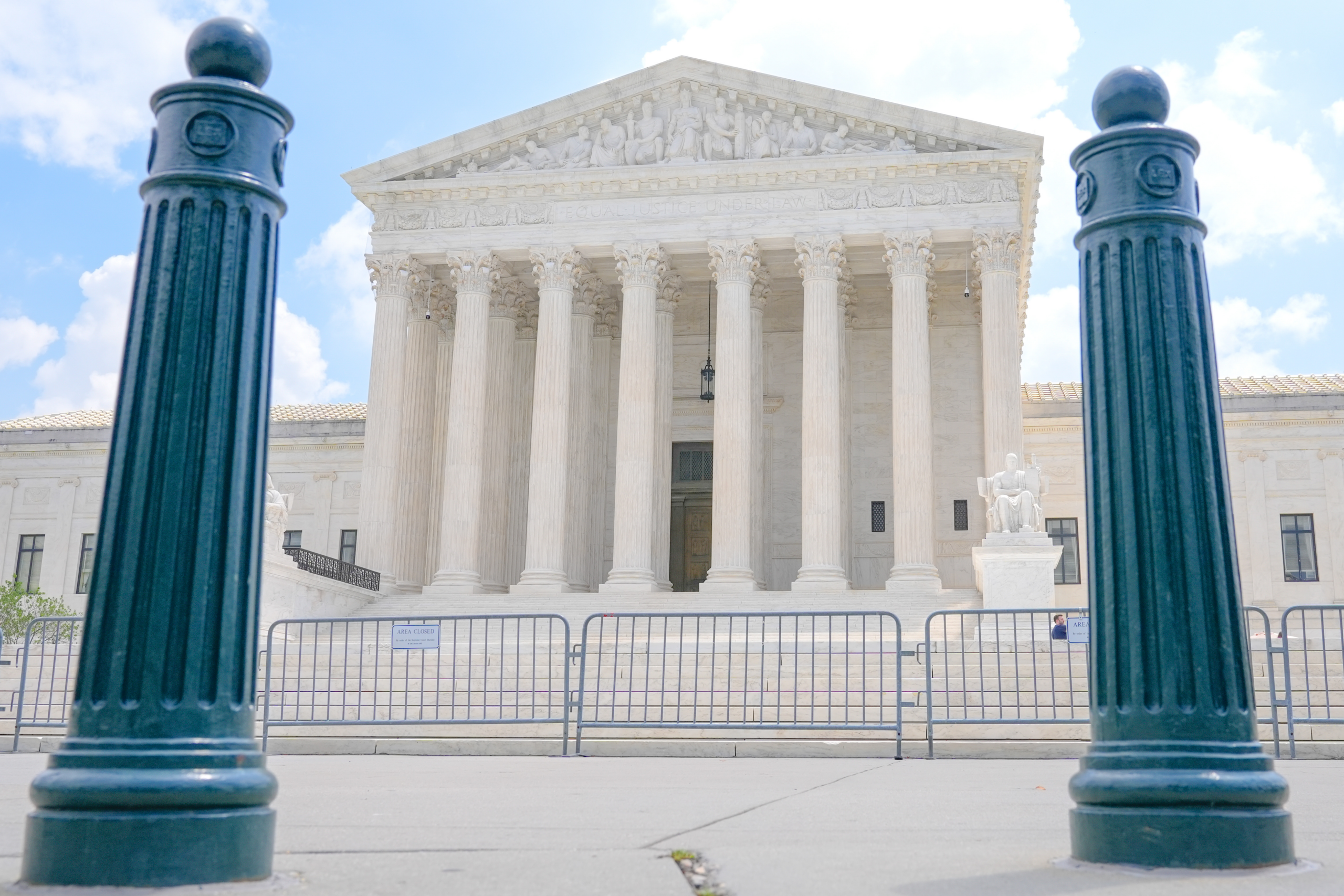 The U.S. Supreme Court building in Washington, D.C., framed between two dark green bollards, with metal barricades and an 'Area Closed' sign across the front steps