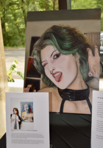 A vibrant painted portrait of Murry Foust with green hair, tongue out, and a playful expression, displayed on an easel at a pop-up gallery alongside an artist statement and smaller photo of Foust.