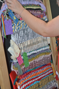 A close-up of a person's hand touching the colorful Art for Murry community loom, with handwritten tags reading "Murry Never Forgotten," "Thank you for sharing your light with the world. You will live on in the community of hearts that you touched. Love you always," and other notes of remembrance woven into the fabric.