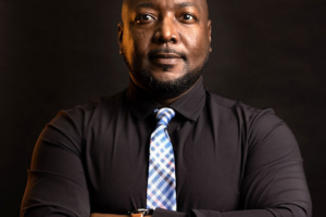 A formal portrait of Kareem Simpson, Covington School Board member and business consultant. He stands with his arms crossed against a dark background, wearing a black button-down shirt and a blue plaid tie. He looks directly into the camera with a composed expression.
