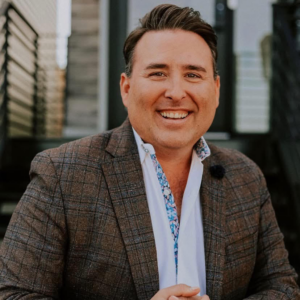 Headshot of J.P. Davis, a white man with dark hair wearing a brown tweed blazer and patterned shirt, smiling outdoors.