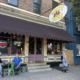 The exterior of Rosie's Tavern in Covington, Kentucky, on a summer afternoon, with patrons sitting outside under a dark awning beneath the bar's oval hanging sign.