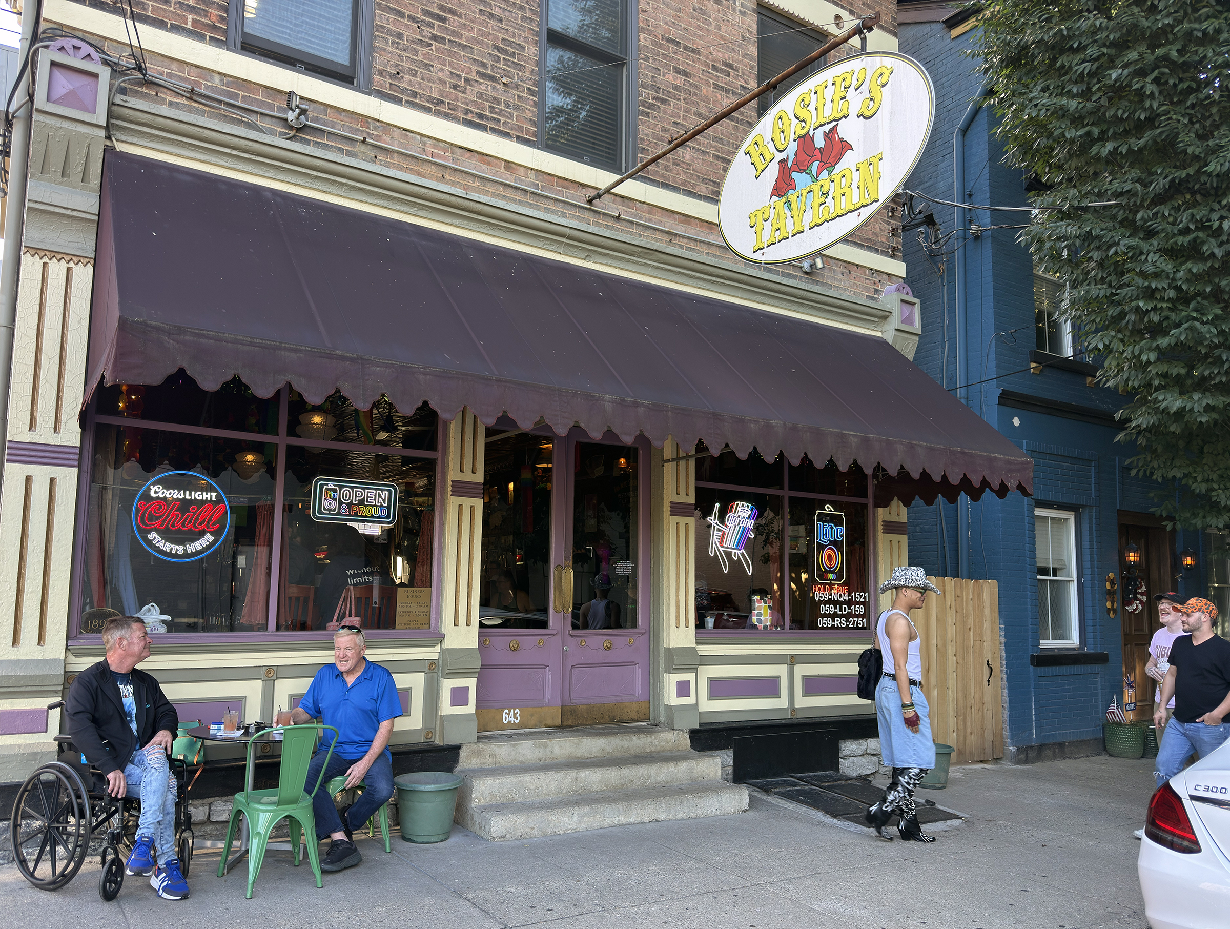 The exterior of Rosie's Tavern in Covington, Kentucky, on a summer afternoon, with patrons sitting outside under a dark awning beneath the bar's oval hanging sign.