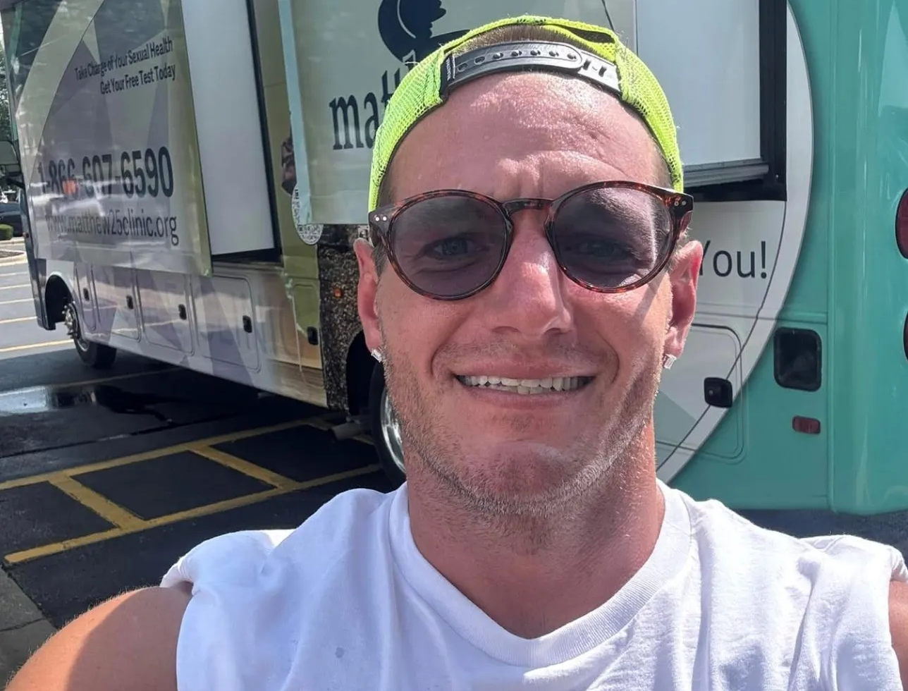 Nathan Wilson smiling in sunglasses and a neon cap in front of a Matthew25 mobile health clinic truck