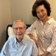 senator-mitch-mcconnell-and-secretary-elaine-chao-07.26.26 Senator Mitch McConnell, seated, smiles alongside his wife, former U.S. Secretary of Transportation Elaine Chao, who stands beside him with her hand on his shoulder in an office setting
