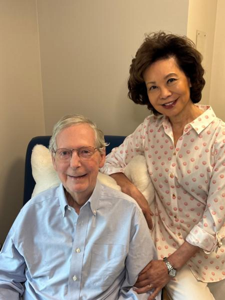 Senator Mitch McConnell, seated, smiles alongside his wife, former U.S. Secretary of Transportation Elaine Chao, who stands beside him with her hand on his shoulder in an office setting