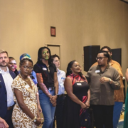 A group of LGBTQ+ community leaders and organizers stand together in a ballroom at the downtown Louisville Marriott during the Creating Change 2027 kickoff event. A person in a lavender blazer speaks into a microphone on the left while the group listens. A presentation screen is partially visible in the background. Attendees wear name tags and represent a diverse range of backgrounds and organizations from across Kentucky and the country.