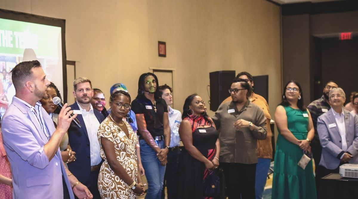 A group of LGBTQ+ community leaders and organizers stand together in a ballroom at the downtown Louisville Marriott during the Creating Change 2027 kickoff event. A person in a lavender blazer speaks into a microphone on the left while the group listens. A presentation screen is partially visible in the background. Attendees wear name tags and represent a diverse range of backgrounds and organizations from across Kentucky and the country.