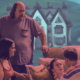 A promotional production photo from Highview Arts Center's staging of Agatha Christie's And Then There Were None. Four cast members are captured mid-scene: a man stands over a sofa looking down, while three other actors engage in tense conversation around it. Behind them, a painted backdrop depicts an isolated manor house surrounded by water — evoking the island setting of Christie's classic mystery. The image is tinted in moody blue and pink tones.