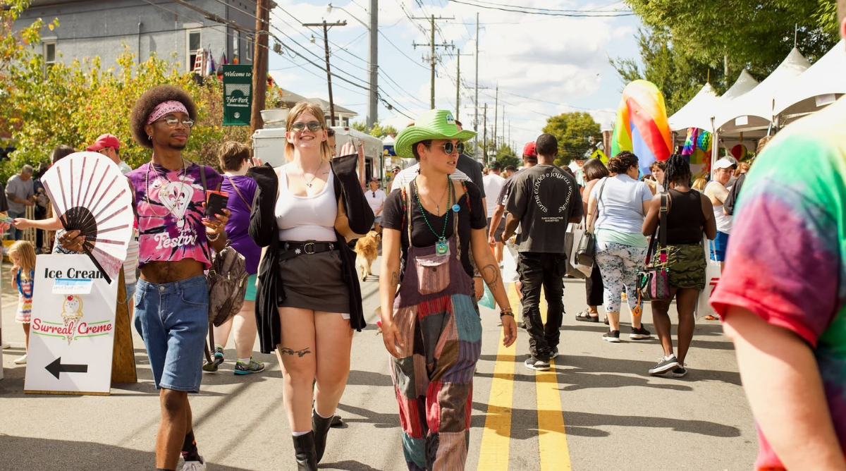 Attendees stroll down a street lined with tents and vendors at a Pride festival in Louisville's Tyler Park neighborhood, passing an ice cream stand and a rainbow-colored art installation