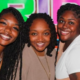 Three smiling Black women pose together in front of a brightly colored Melanin Pride Weekend banner in Louisville.