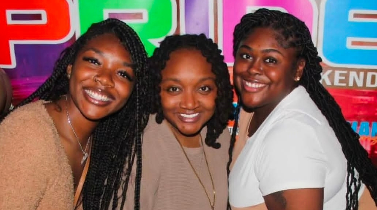 Three smiling Black women pose together in front of a brightly colored Melanin Pride Weekend banner in Louisville.