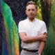 Kentucky Poet Laureate Silas House stands with arms crossed in front of a composite background featuring a rainbow streak and lush green trees.