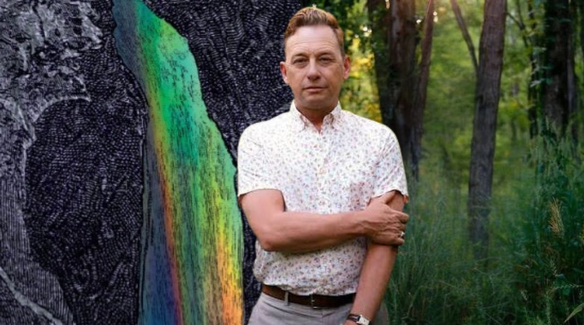 Kentucky Poet Laureate Silas House stands with arms crossed in front of a composite background featuring a rainbow streak and lush green trees.