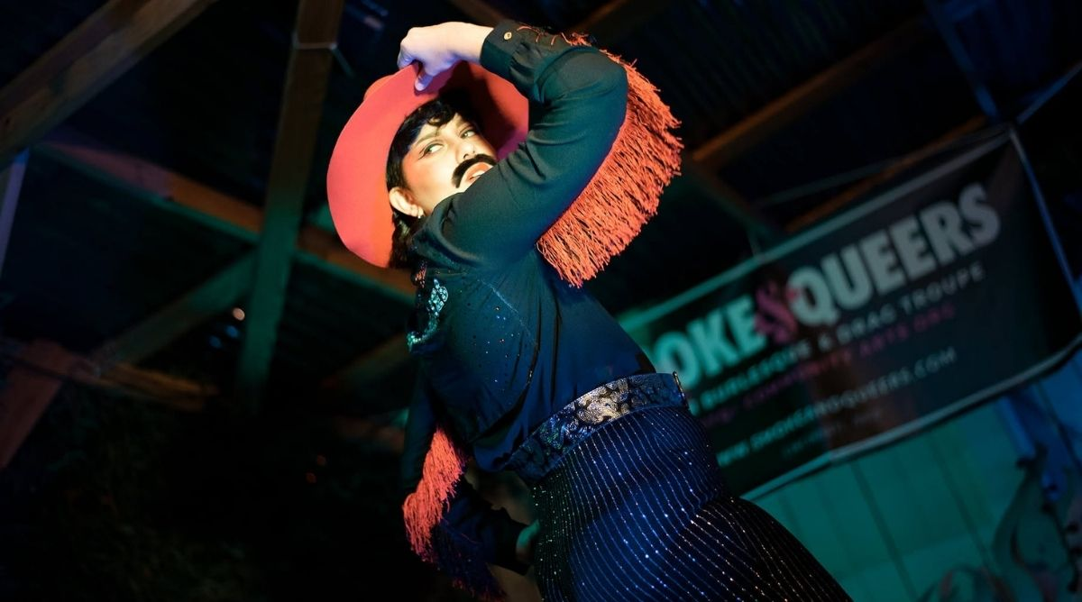 Seymour Hiney performs at a Smoke & Queers queer burlesque show in a sequined outfit with red fringe, arms raised on stage.