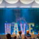 Guests mingle at a previous WAVE Foundation Make WAVEs Gala at Newport Aquarium, with a large illuminated WAVE sign in front of a floor-to-ceiling aquarium tank.