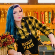 Mercury Stardust leans over a wooden table with a toolkit, holding a hammer, and wearing an apron that say "HANDY MA'AM."